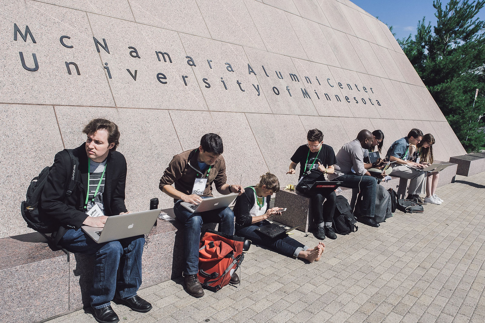 A row of Source-con participants sit on or near benches in front of the McNamara Alumni Center, the conference venue, to co-work.