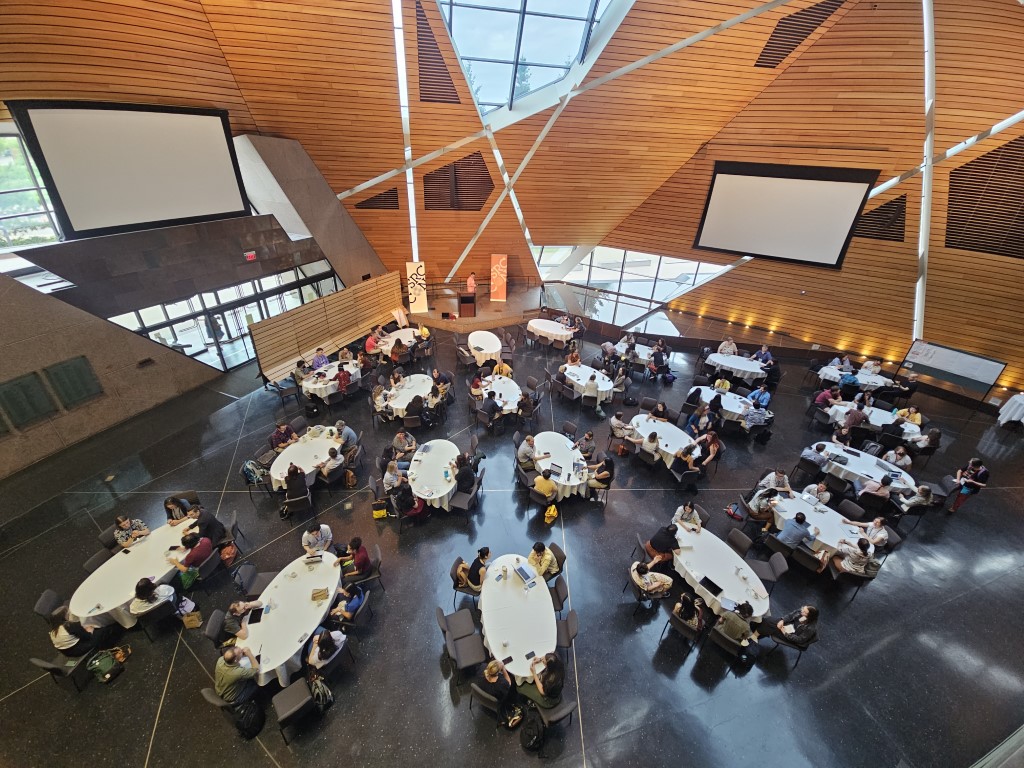An overhead view of Memorial Hall at Source-con 2025 in Minneapolis with conference attendees sitting around tables under the skylights of the main seating area in Memorial Hall.